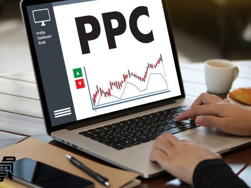 The image shows a person using a laptop with a screen displaying the term "PPC" in large, bold letters. Beneath it, there is a line graph with upward and downward arrows indicating trends or performance metrics. On the left side of the screen, there is a sidebar menu with options labeled "Profile," "Dashboard," and "Email." The person is typing on the keyboard of the laptop. In the bottom left corner of the image, there's an icon resembling a computer monitor with the word "ADS" written inside it, representing the concept of online advertising.