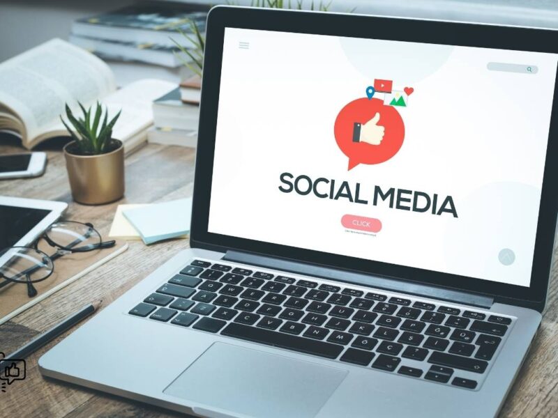 A laptop is open on a wooden desk, with the screen displaying a "Social Media" interface that features a large red speech bubble with a thumbs-up icon inside it. In the background, there are open books, a notebook, and a pair of glasses suggesting a study or work environment.