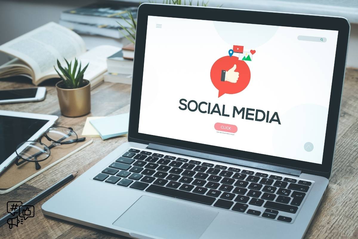 A laptop is open on a wooden desk, with the screen displaying a "Social Media" interface that features a large red speech bubble with a thumbs-up icon inside it. In the background, there are open books, a notebook, and a pair of glasses suggesting a study or work environment.